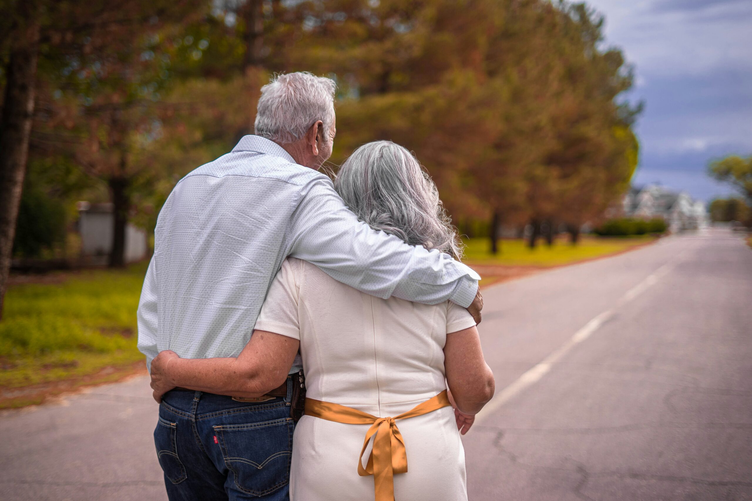 An image of a senior couple walking down the road with their backs to the camera. Their arms are wrapped around each other. The road is lined with trees that have autumn foliage, and the sky is stormy, but the couple is embracing as they walk.