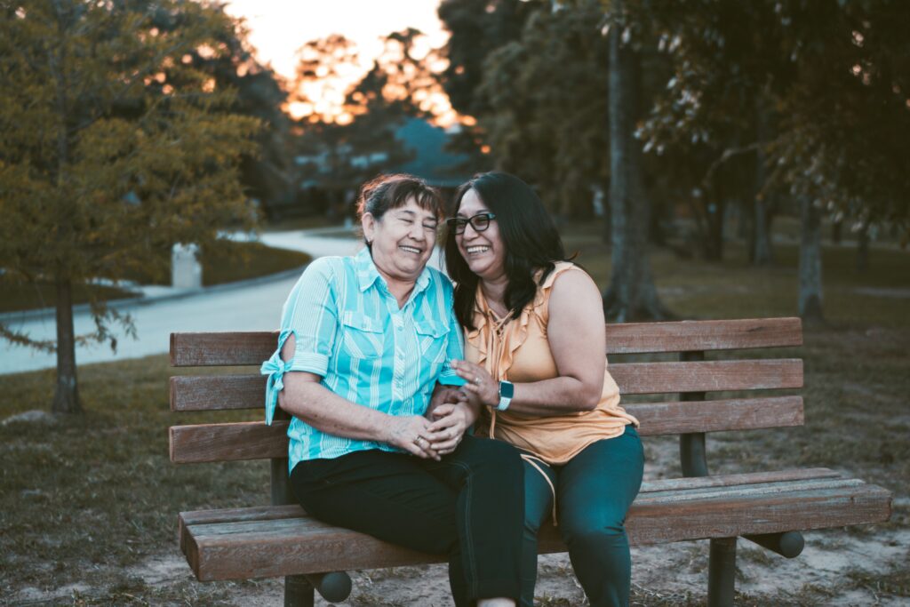 Two women, one senior and one younger, smiling and sitting on a park bench, sharing a moment of joy in an outdoor park setting.